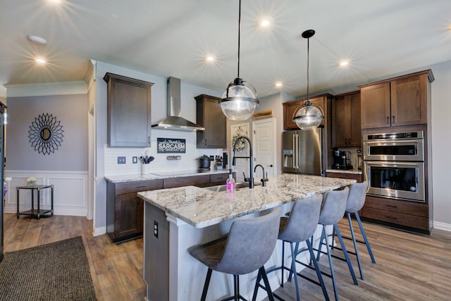 kitchen with a sink, a breakfast bar area, wall chimney range hood, appliances with stainless steel finishes, and wood finished floors