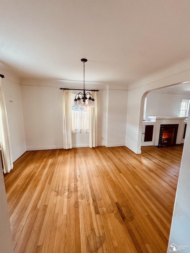unfurnished dining area featuring light wood-style flooring, arched walkways, baseboards, a chandelier, and a lit fireplace