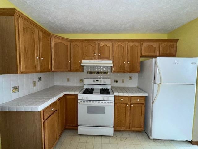 kitchen featuring tile counters, tasteful backsplash, brown cabinetry, under cabinet range hood, and white appliances