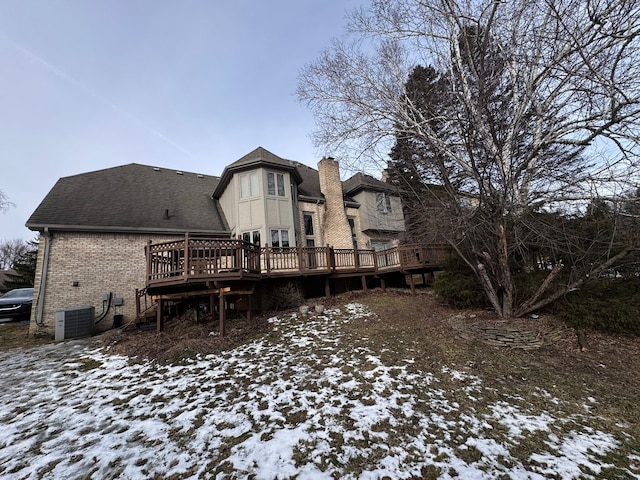snow covered back of property featuring central AC, a deck, a chimney, brick siding, and roof with shingles