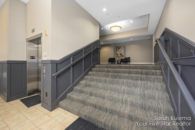 stairway featuring elevator, a decorative wall, tile patterned floors, and wainscoting