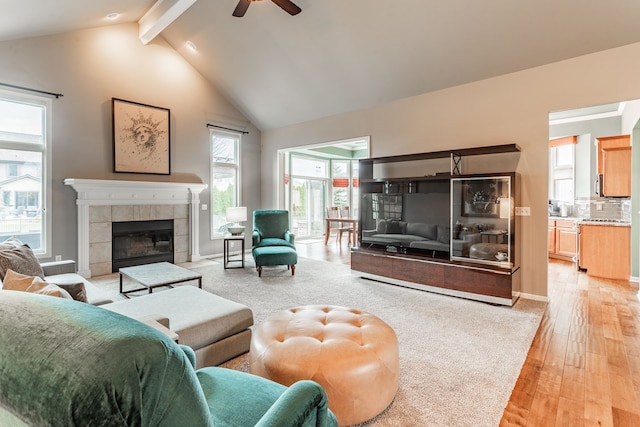 living area with a tile fireplace, beam ceiling, light wood-style floors, high vaulted ceiling, and a ceiling fan
