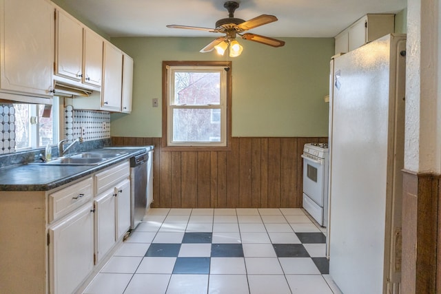 kitchen with a wainscoted wall, wooden walls, white appliances, dark countertops, and a sink
