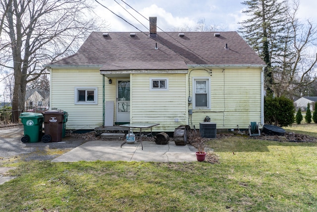 rear view of property featuring a shingled roof, a patio, a lawn, central AC, and a chimney