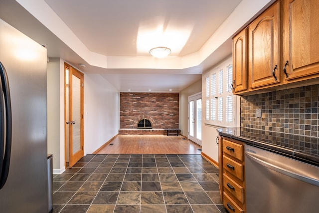 kitchen with a brick fireplace, brown cabinets, appliances with stainless steel finishes, a tray ceiling, and backsplash