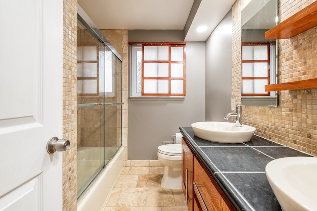 full bath featuring stone tile floors, toilet, a sink, bath / shower combo with glass door, and double vanity