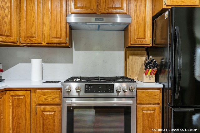 kitchen with stainless steel gas range oven, ventilation hood, brown cabinetry, and freestanding refrigerator
