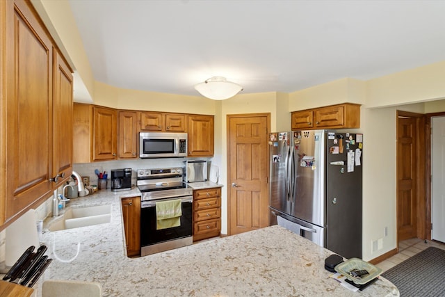 kitchen featuring appliances with stainless steel finishes, brown cabinets, a sink, light stone counters, and a peninsula