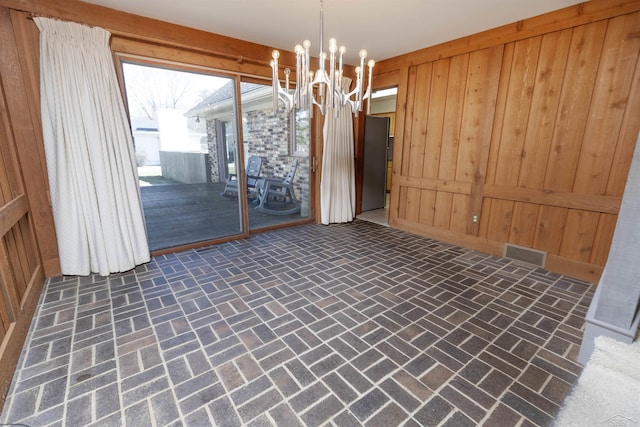 unfurnished dining area featuring brick floor, visible vents, an inviting chandelier, and wood walls