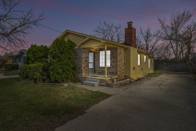 property exterior at dusk featuring concrete driveway, a chimney, brick siding, a yard, and fence