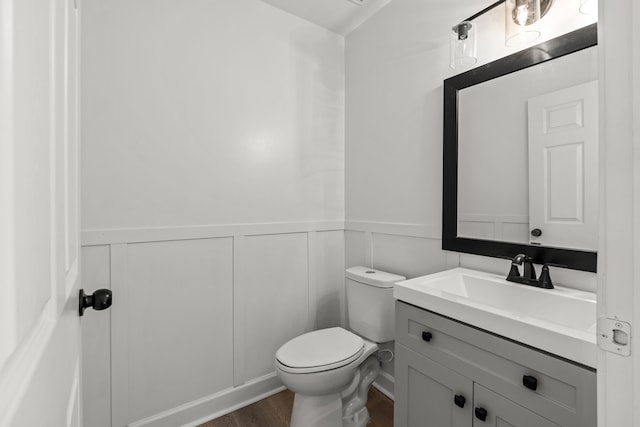 bathroom featuring a wainscoted wall, toilet, vanity, wood finished floors, and a decorative wall