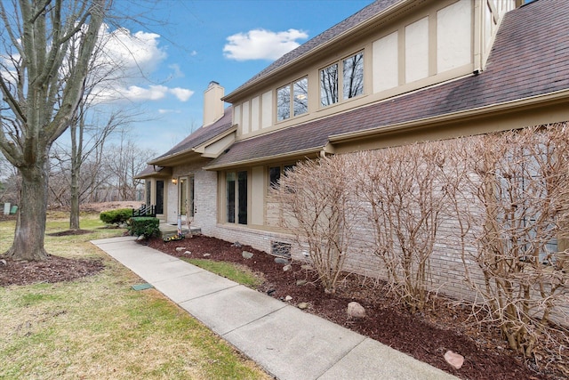 view of side of home with a chimney, stucco siding, brick siding, roof with shingles, and a lawn