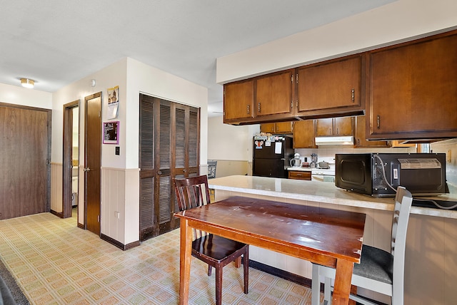 kitchen with a wainscoted wall, light countertops, under cabinet range hood, black appliances, and brown cabinetry