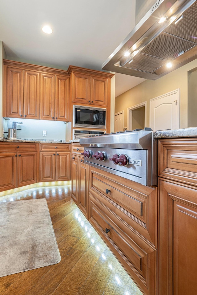 kitchen with stainless steel stovetop, wood finished floors, brown cabinets, black microwave, and wall chimney range hood