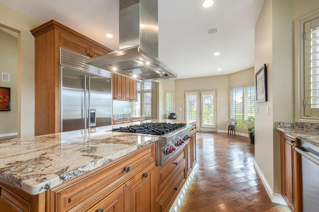 kitchen with appliances with stainless steel finishes, light stone countertops, island exhaust hood, and brown cabinets