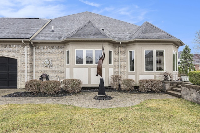 exterior space with stucco siding, roof with shingles, an attached garage, a front lawn, and brick siding