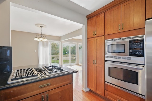 kitchen with appliances with stainless steel finishes, brown cabinetry, dark countertops, light wood finished floors, and a chandelier