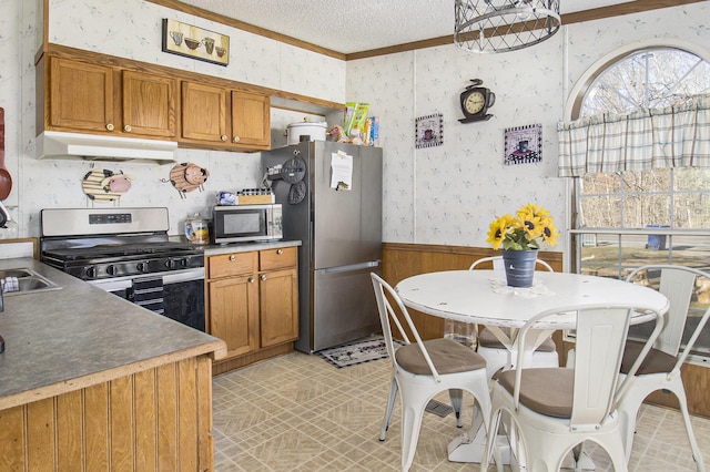 kitchen featuring a wainscoted wall, appliances with stainless steel finishes, a textured ceiling, under cabinet range hood, and wallpapered walls