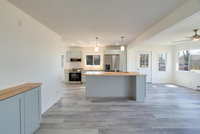 kitchen featuring light wood finished floors, a kitchen island with sink, butcher block counters, appliances with stainless steel finishes, and hanging light fixtures