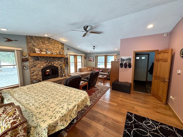 bedroom featuring a textured ceiling, ceiling fan, a stone fireplace, light wood-style floors, and vaulted ceiling
