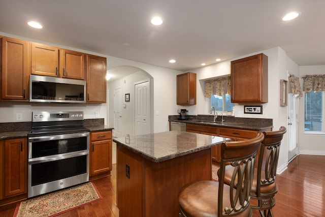 kitchen with a sink, dark wood finished floors, arched walkways, a center island, and appliances with stainless steel finishes