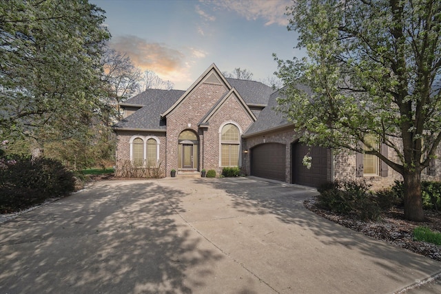 french country inspired facade featuring a shingled roof, an attached garage, driveway, and brick siding