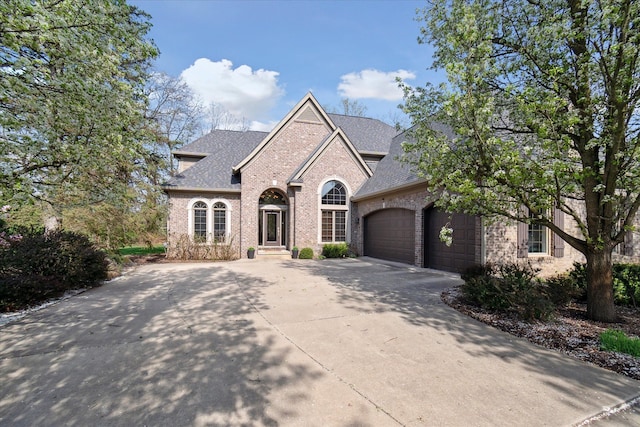 french country home featuring a garage, driveway, roof with shingles, and brick siding