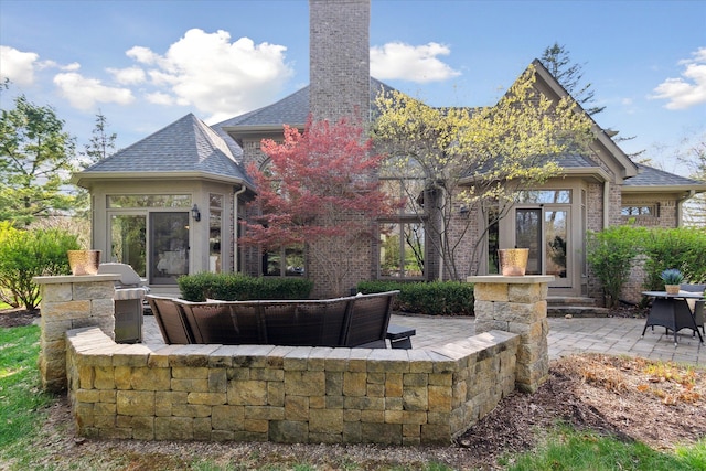 rear view of property with a shingled roof, a patio, a chimney, and brick siding