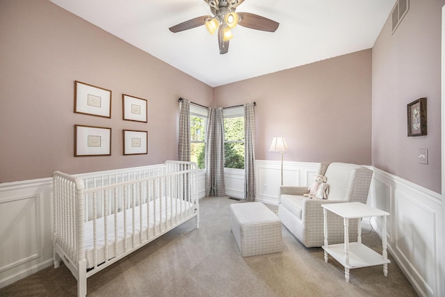 carpeted bedroom with a nursery area, a ceiling fan, visible vents, and a wainscoted wall
