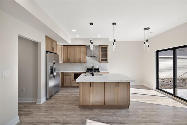 kitchen featuring baseboards, wall chimney exhaust hood, appliances with stainless steel finishes, a kitchen island with sink, and light wood-style flooring