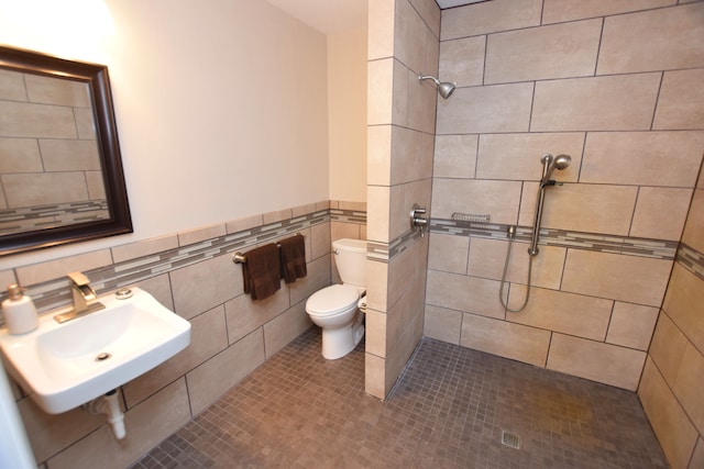 bathroom with a tile shower, a wainscoted wall, toilet, a sink, and tile walls