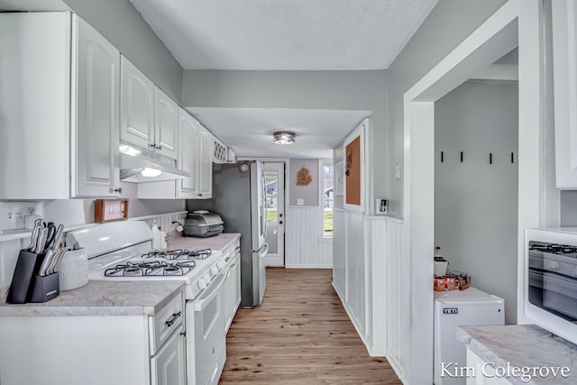 kitchen with white gas stove, white cabinetry, light countertops, under cabinet range hood, and wainscoting