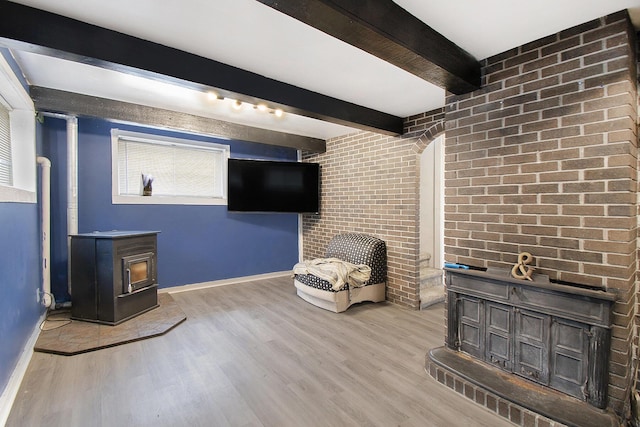 unfurnished room featuring baseboards, brick wall, a wood stove, beamed ceiling, and wood finished floors