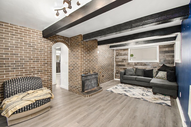 living area featuring arched walkways, brick wall, a fireplace, beamed ceiling, and wood finished floors