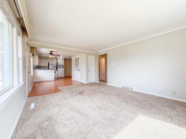 unfurnished living room with crown molding, ceiling fan, light colored carpet, baseboards, and visible vents