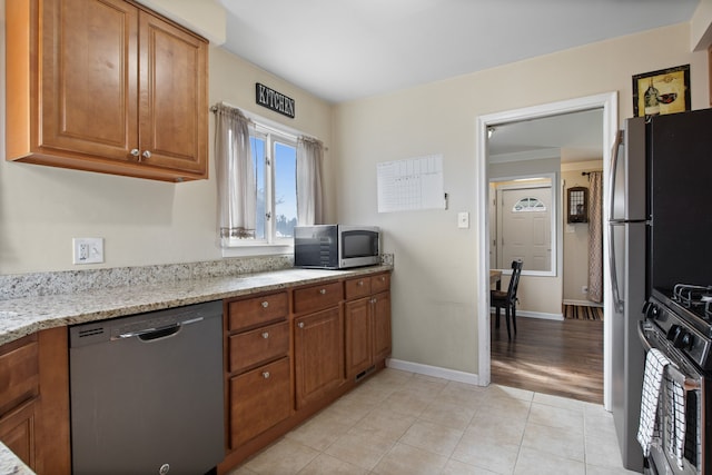 kitchen featuring light tile patterned floors, baseboards, appliances with stainless steel finishes, brown cabinetry, and light stone counters