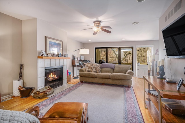 living area featuring a tiled fireplace, visible vents, a ceiling fan, and wood finished floors