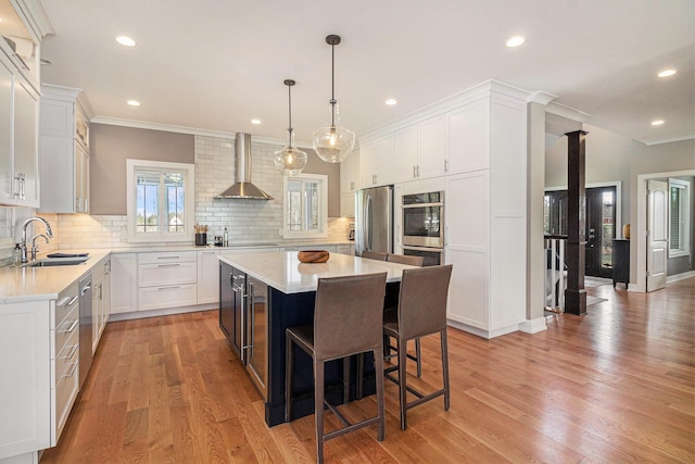 kitchen featuring wall chimney exhaust hood, a center island, appliances with stainless steel finishes, light countertops, and a sink