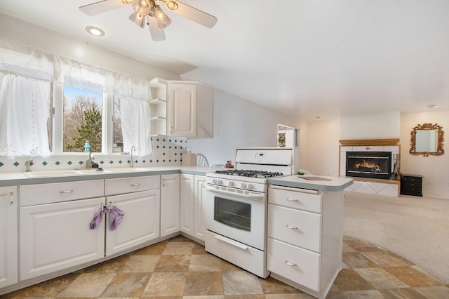 kitchen featuring white range with gas cooktop, a peninsula, light colored carpet, a ceiling fan, and a tile fireplace