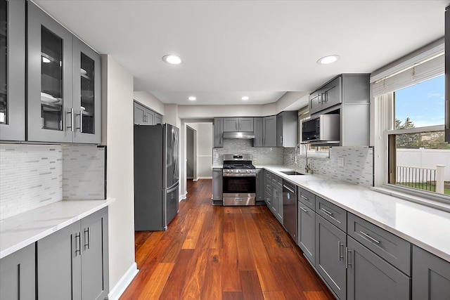 kitchen with gray cabinets, light stone countertops, under cabinet range hood, appliances with stainless steel finishes, and a sink