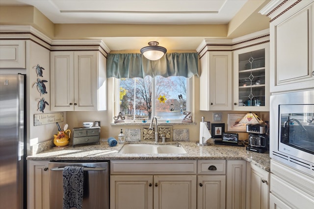 kitchen featuring glass insert cabinets, light stone counters, a sink, stainless steel appliances, and cream cabinets
