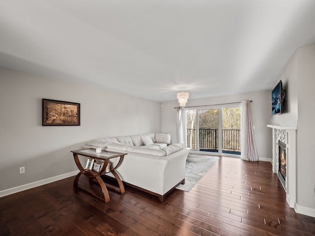 living room featuring a notable chandelier, dark wood finished floors, a glass covered fireplace, and baseboards