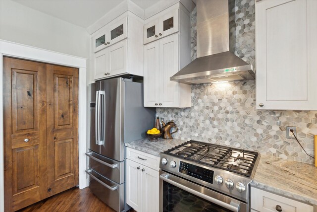 kitchen featuring appliances with stainless steel finishes, wall chimney exhaust hood, light stone counters, and glass insert cabinets