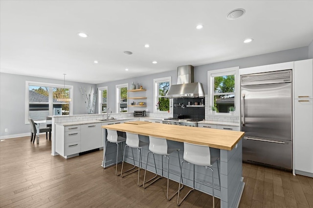 kitchen featuring open shelves, wall chimney exhaust hood, a wealth of natural light, built in refrigerator, and butcher block counters