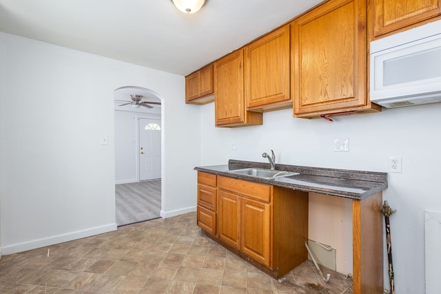 kitchen with a sink, brown cabinetry, ceiling fan, white microwave, and baseboards