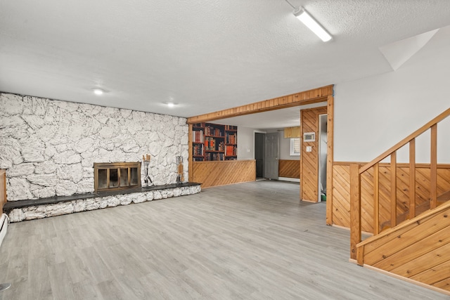 unfurnished living room with a textured ceiling, a stone fireplace, wood finished floors, a wainscoted wall, and wooden walls