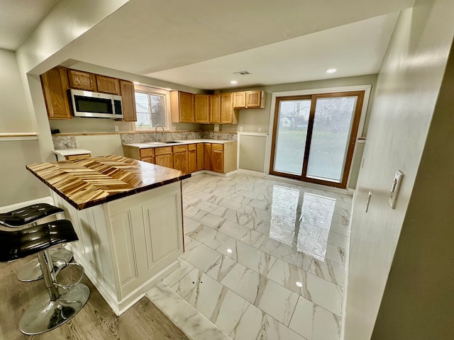 kitchen featuring baseboards, brown cabinetry, stainless steel microwave, recessed lighting, and marble finish floor