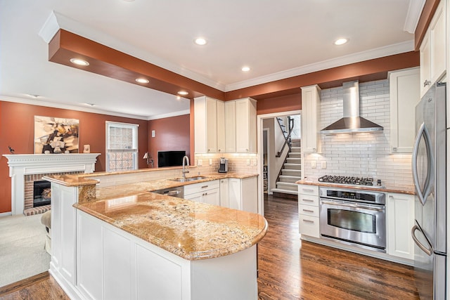 kitchen with a sink, stainless steel appliances, a fireplace, a peninsula, and wall chimney range hood