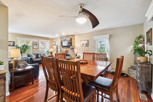 dining space featuring a textured ceiling, ceiling fan, a stone fireplace, and wood finished floors