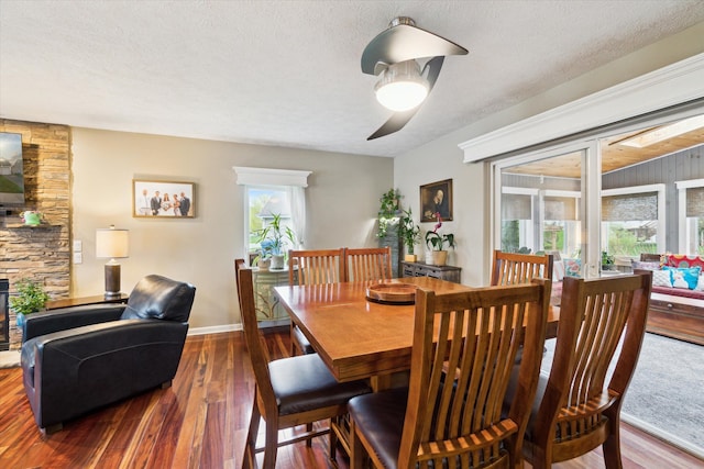 dining room featuring ceiling fan, a textured ceiling, a stone fireplace, baseboards, and wood finished floors